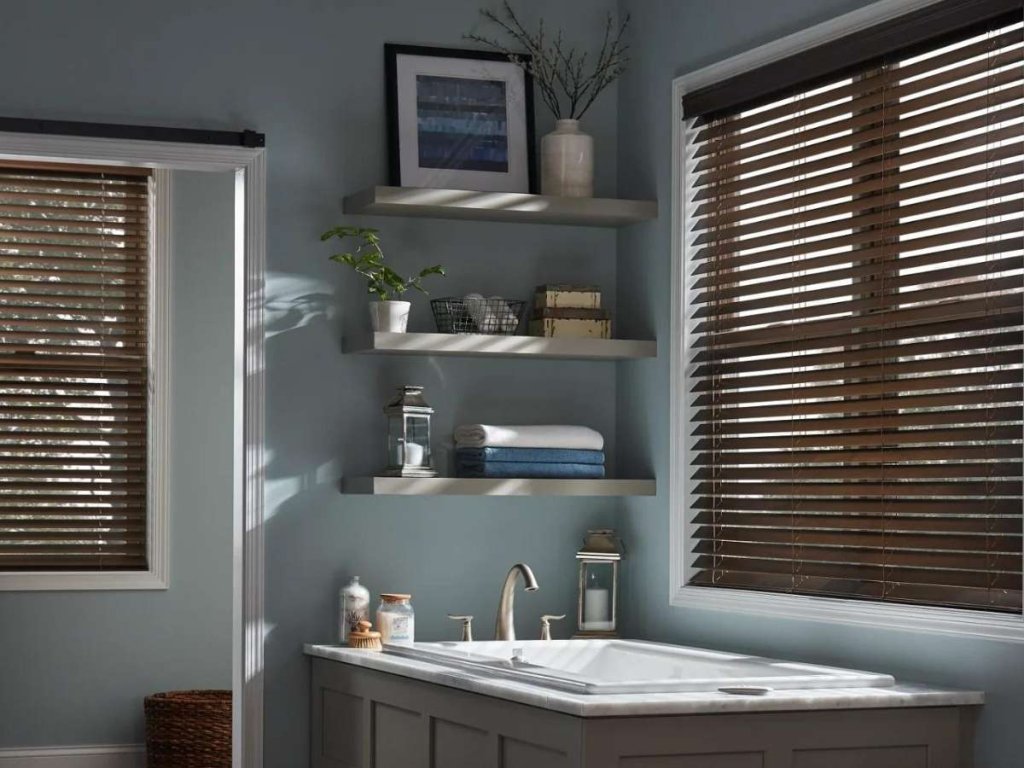 A bathroom with light blue walls, featuring a bathtub beneath two windows with wooden blinds. Floating shelves above the bathtub hold plants, towels, and decor items.